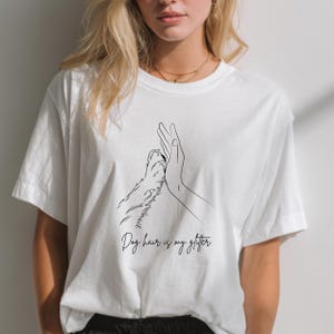 Könnte beinhalten: Weißes T-Shirt mit einer schwarzen Strichzeichnung einer Hundepfote, die eine Hand berührt. Der Text "Dog hair is my glitter" ist in einer Schreibschrift unter der Illustration geschrieben. Ein lässiger Alltagsartikel.