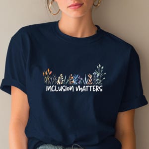 Könnte beinhalten: Marineblaues T-Shirt mit dem weißen Schriftzug "INCLUSION MATTERS" über einem farbenfrohen Blumendesign. Das Design enthält orange, gelbe, violette und grüne Elemente. Das T-Shirt hat kurze Ärmel.