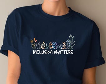 May include: Navy blue t-shirt with the text "INCLUSION MATTERS" in white, beneath a colorful floral design. The design includes orange, yellow, purple, and green elements. The shirt has short sleeves.