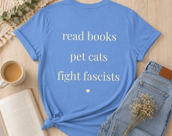 May include: A light blue short-sleeved t-shirt featuring the cream-colored text "read books", "pet cats", and "fight fascists" stacked vertically, followed by a small cream-colored heart symbol centered below the text on the front of the shirt.