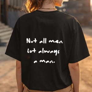 Könnte beinhalten: Schwarzes T-Shirt mit weißem Text: „Not all men but always a man.“ Das Shirt ist ein lässiger Stil, wahrscheinlich aus Baumwolle oder einem ähnlichen Stoff. Der Text ist handschriftlich.