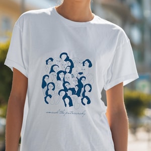 Puede incluir: Camiseta blanca con una ilustración azul de múltiples rostros y figuras. El texto "smash the patriarchy" está escrito en cursiva debajo de la ilustración. La camiseta tiene mangas cortas y cuello redondo.