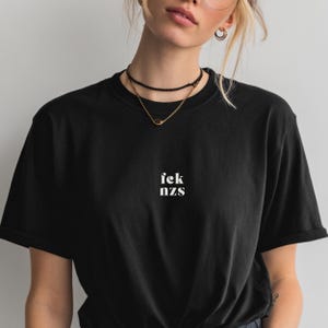 Könnte beinhalten: Schwarzes T-Shirt mit dem weißen Text "fek nzs" mittig auf der Brust. Das Shirt hat einen Rundhalsausschnitt und kurze Ärmel. Das Model trägt einen schwarzen Choker und eine goldene Halskette.