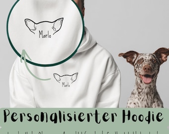 May include: White hoodie with a black outline of dog ears and the name "Marlo" printed below. The hoodie has the text "Personalisierter Hoodie" and other text in German. A dog with brown and white spots is in the background.