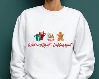 May include: White sweatshirt featuring Christmas-themed graphics: a green mug with a Christmas tree, a red mug with a drink, a snow globe with a house, and a gingerbread man. The text "Weihnachtszeit-Lieblingszeit" is written in red cursive.
