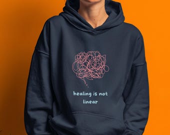 May include: Navy blue hoodie with a pink, scribbled brain graphic and the text "healing is not linear". The hoodie has a drawstring hood and a front pocket. The background is orange.