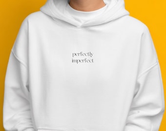 May include: A white hooded sweatshirt with the words "perfectly imperfect" printed in black. The sweatshirt has a drawstring hood and a kangaroo pocket. The background is a solid yellow.