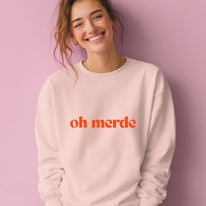 Könnte beinhalten: Hellrosa Sweatshirt mit dem orangefarbenen Schriftzug "oh merde". Das Sweatshirt mit Rundhalsausschnitt scheint aus einem weichen Material zu sein. Das Bild zeigt eine Person, die das Sweatshirt trägt.