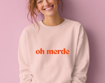 May include: A light pink sweatshirt with the phrase "oh merde" in orange lettering. The sweatshirt is a crew neck style and appears to be made of a soft material. The image is of a person wearing the sweatshirt.