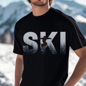 Op de afbeelding: Zwart T-shirt met een witte afbeelding van het woord "SKI" met een silhouet van een skiër die in het midden van de letters springt. De achtergrond van de afbeelding is een besneeuwd bos.