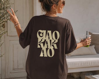 May include: A dark brown short-sleeve t-shirt featuring the text "CIAO KAKAO" printed in a large, cream-colored, wavy, retro-style font on the back. The shirt has a crew neckline and a loose, relaxed fit, showcasing the graphic design clearly.
