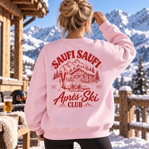 Puede incluir: Una sudadera de manga larga en color rosa claro con un gráfico rojo en la espalda. El diseño incluye una cabaña de montaña, esquís, jarras de cerveza y el texto "SAUFI SAUFI APRÈS-SKI CLUB EST. 2025" con "APRÈS SKI" en el tejado.