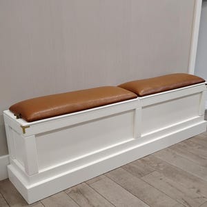 May include: A white storage bench with two brown leather cushions. The bench has a rectangular shape and is made of wood. The cushions are rectangular and sit atop the bench. The bench is placed against a light gray wall.