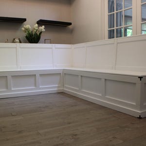 Könnte beinhalten: Eine große, weiße Eckbank mit einem Paneeldesign. Die Bank ist aus Holz gefertigt und hat eine rechteckige Form mit flacher Sitzfläche und Rückenlehne. Die Bank befindet sich in einem Raum mit Holzboden und einer neutralfarbenen Wand.