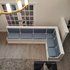 May include: An L-shaped corner bench featuring a white wooden frame and smooth, light blue upholstered seat and back cushions. The bench is positioned against a wall, providing integrated seating with a clean, structured design.
