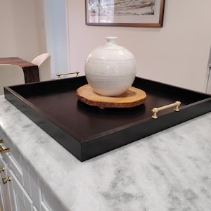 May include: A dark brown wooden serving tray with gold handles. A white ceramic vase with a textured pattern sits on a wooden slice in the center of the tray. The tray is on a marble countertop.