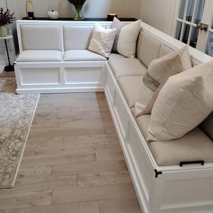 May include: An L-shaped white wooden corner bench with light beige upholstered seat and back cushions. The bench features a paneled base and is decorated with several throw pillows in varying shades of cream, beige, and textured white fabric.