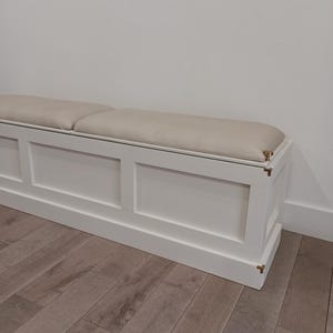 May include: A white rectangular storage bench featuring a smooth, light beige upholstered seat cushion. The base is constructed with clean, paneled sides and includes small, metallic gold-toned corner accents at the edges of the frame.