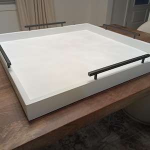 May include: A large, square white serving tray with black handles. The tray is made of wood and sits on a dark brown wooden table. The handles are rectangular and are attached to the sides of the tray. The tray is empty.