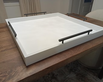 May include: A large, square white serving tray with black handles. The tray is made of wood and sits on a dark brown wooden table. The handles are rectangular and are attached to the sides of the tray. The tray is empty.