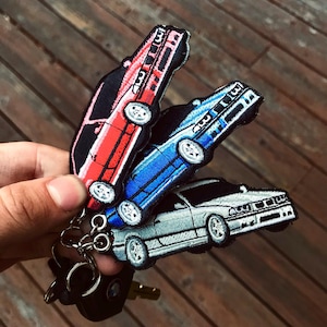 May include: Three car keychains, one red, one blue, and one silver, are attached to a silver key ring. The keychains are made of fabric and feature embroidered car designs.