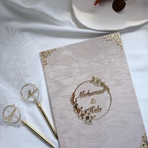Puede incluir: Una invitación de boda color rubor con detalles florales dorados y los nombres "Mohamad & Hala" en un diseño circular dorado. También se ven dos bolígrafos dorados con el nombre "Hala".