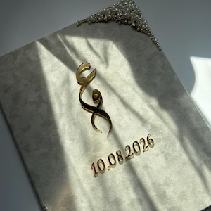 May include: A rectangular beige velvet-textured board decorated with white pearls in two corners. The centre features a gold-coloured Arabic letter and the date 10.08.2026 in gold-coloured lettering. The surface has a soft, matte finish.