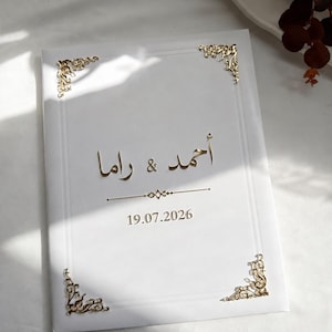 Puede incluir: Una tarjeta de invitación rectangular blanca con detalles de filigrana metálica dorada en las esquinas. El centro muestra el texto en árabe "أحمد & راما" en letras doradas, seguido de una línea horizontal y la fecha "19.07.2026" en oro.