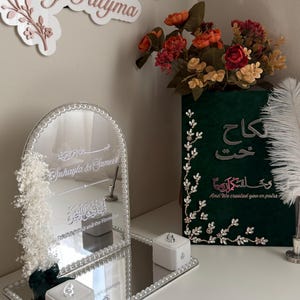 May include: A rectangular mirrored tray with a beaded border, featuring an arched mirror with the text "Suhayla & Sameer" and "And We created you in pairs". Two white ring boxes sit on the tray, accompanied by a dark green sign with Arabic text.