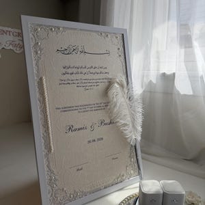 Könnte beinhalten: Eine weiß gerahmte Heiratsurkunde auf cremefarbenem Papier mit Spitzenbordüren, Perlenakzenten und einem weißen Federkiel. Das Dokument enthält arabische Kalligrafie, englischen Text, die Namen Ramiz & Bushra sowie das Datum 20.08.2026.