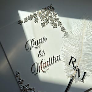 May include: A white rectangular guest book featuring the silver text Ryan & Madiha, decorated with clear crystal embellishments in the corners. A white feather quill pen with a silver R&M topper rests on the surface of the book.