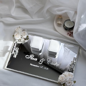 Puede incluir: Una bandeja rectangular de espejo con borde de perlas, que incluye dos cajas blancas para anillos, asas plateadas y detalles florales blancos. La superficie está grabada con el texto "Ilza & Costa" y la fecha "November 14th, 2026" en blanco.