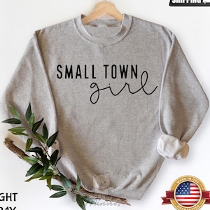 May include: Light gray sweatshirt with the text "SMALL TOWN girl" in black script. The sweatshirt is displayed on a wooden hanger. The image also includes a sprig of greenery and a "Made in USA" sticker.