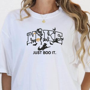 May include: White t-shirt with a black graphic that says "NIKE" with Halloween-themed imagery and the text "JUST BOO IT." The model is wearing black and white checkered shorts. A "NEXT DAY SHIPPING" sticker is in the top right corner.