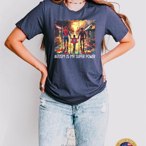 May include: A navy blue t-shirt with a graphic of three superheroes walking on a puzzle piece sidewalk. The text "AUTISM IS MY SUPER POWER" is printed below the graphic.