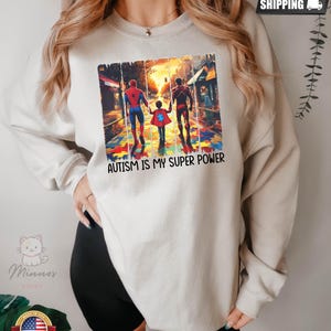 May include: A white sweatshirt with a graphic of two superheroes, Spiderman and Iron Man, walking with a child on a street with a puzzle piece background. The text "Autism is my super power" is printed below the graphic.