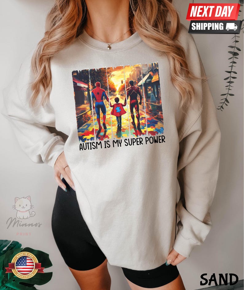 May include: A white sweatshirt with a graphic of two superheroes, Spiderman and Iron Man, walking with a child on a street with a puzzle piece background. The text "Autism is my super power" is printed below the graphic.