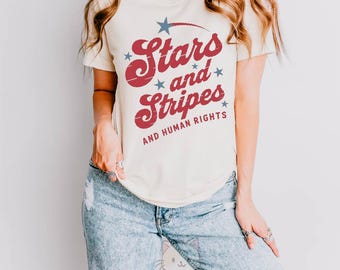 May include: Cream-colored t-shirt with the text "Stars and Stripes AND HUMAN RIGHTS" in red and blue. The shirt features a star graphic and is worn with light-wash ripped jeans. The image also includes a "Made in USA" seal.