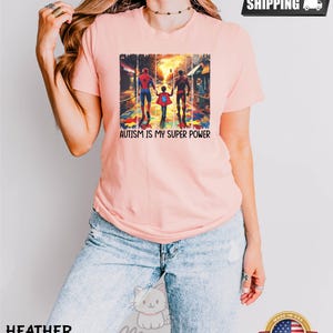 May include: A pink t-shirt with a graphic of three superheroes walking on a street with a puzzle piece pattern. The text "AUTISM IS MY SUPER POWER" is printed below the graphic.