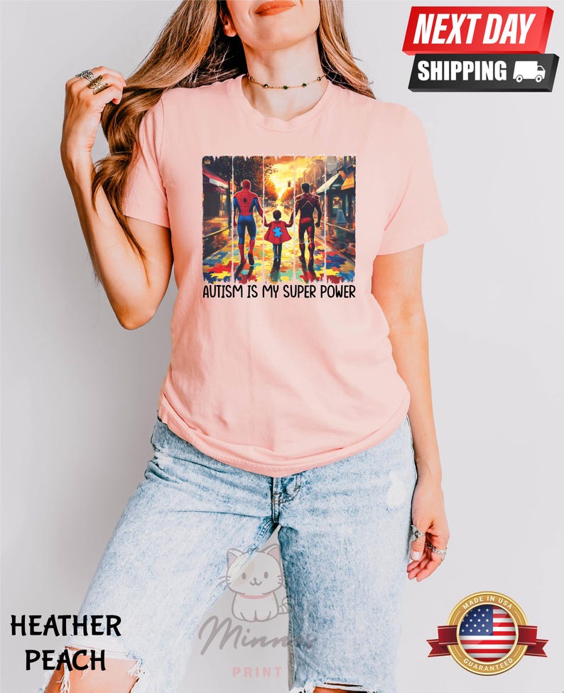 May include: A pink t-shirt with a graphic of three superheroes walking on a street with a puzzle piece pattern. The text "AUTISM IS MY SUPER POWER" is printed below the graphic.