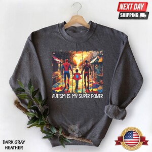 May include: Dark gray heather sweatshirt with a graphic of three figures walking on a path made of puzzle pieces. The figures are wearing superhero costumes. The text "AUTISM IS MY SUPER POWER" is printed below the graphic.