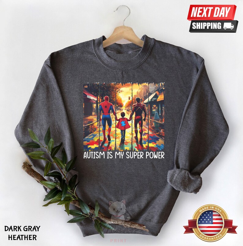 May include: Dark gray heather sweatshirt with a graphic of three figures walking on a path made of puzzle pieces. The figures are wearing superhero costumes. The text "AUTISM IS MY SUPER POWER" is printed below the graphic.