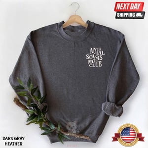 A gray sweatshirt with the text "ANTI SOCIAL MOMS CLUB" printed on it, hanging on a wooden hanger. The sweatshirt is displayed against a white background, with a small plant branch and a "NEXT DAY SHIPPING" sign visible.