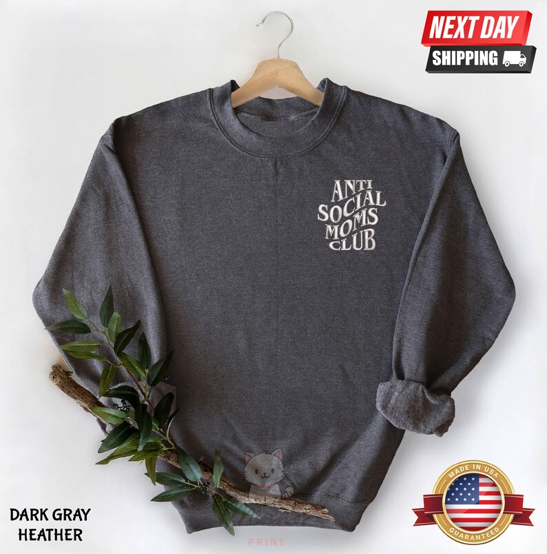 A gray sweatshirt with the text "ANTI SOCIAL MOMS CLUB" printed on it, hanging on a wooden hanger. The sweatshirt is displayed against a white background, with a small plant branch and a "NEXT DAY SHIPPING" sign visible.