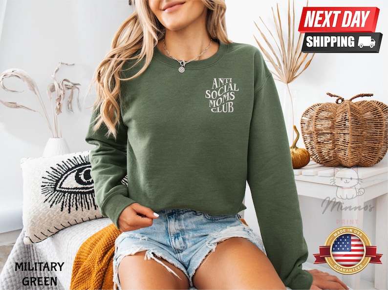 A woman wearing a green sweatshirt with the text "ANT SOCIAL MOMS CLUB" printed on it, paired with denim shorts. She is sitting on a couch with a woven basket nearby.