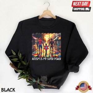 May include: Black sweatshirt with a graphic of three figures walking on a street with a puzzle piece pattern on the ground. The figures are wearing superhero costumes. The text "Autism is my super power" is printed below the graphic.