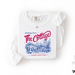 May include: White long-sleeve shirt with a graphic that says "Greetings From The Cottage" in pink and blue. The graphic features a cottage illustration and the text "Lanaudière, Québec, Canada". A silver necklace is visible.