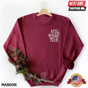 A maroon sweatshirt with the text "ANTI SOCIAL MOMS CLUB" printed on it, hanging on a wooden hanger. The sweatshirt also features a small cat design.