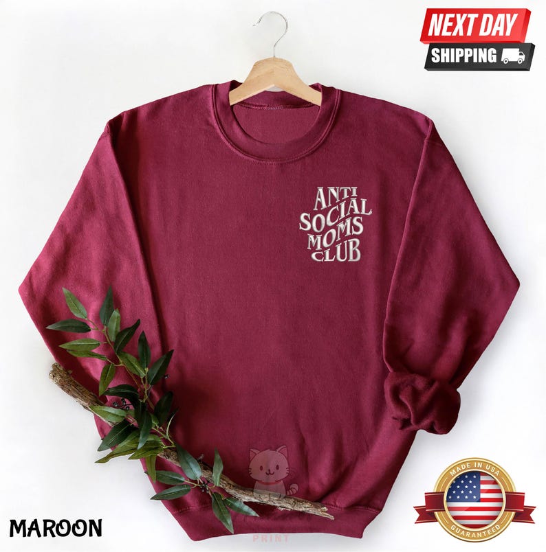A maroon sweatshirt with the text "ANTI SOCIAL MOMS CLUB" printed on it, hanging on a wooden hanger. The sweatshirt also features a small cat design.