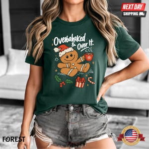 May include: Forest green t-shirt with a gingerbread man graphic wearing a Santa hat and holding a mug. The text "Overbaked & Over It" is above the gingerbread man. Christmas-themed design with holly, a gift, and a candy cane.